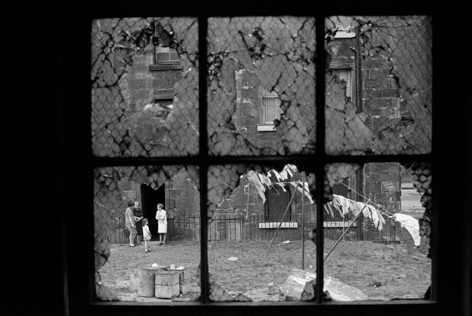 #60 Looking out through broken windows onto a courtyard scene with laundry and four residents of slum housing in the notorious Gorbals district of Glasgow, 1969.