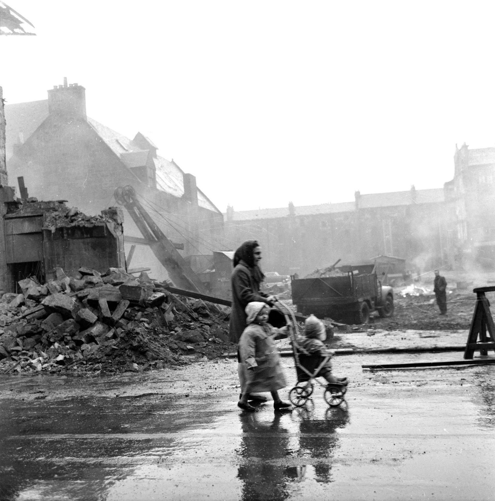 #76 A woman with two young children walking past a demolition site in the Gorbals area of Glasgow, 1962