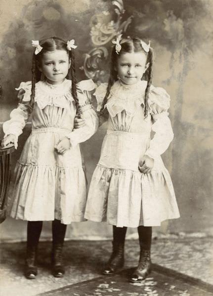 #94 Twins Emma (1893-1974) and Marge (1893-1973) Willms of Milwaukee, Wisconsin standing in front of a painted backdrop, 1898