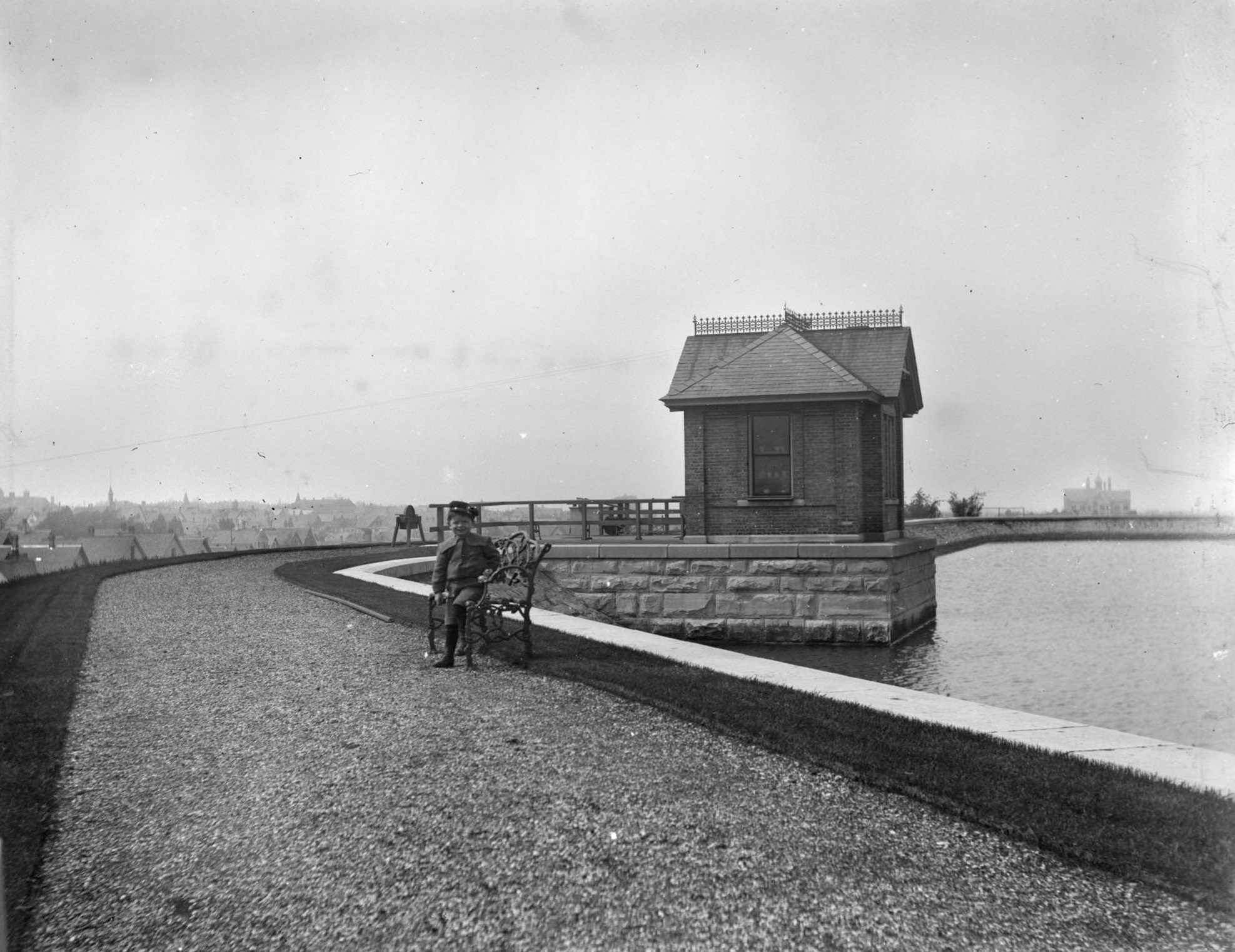 #110 View down gravel path towards Syl seated on a bench along the edge of a reservoir on a hill, Milwaukee, Wisconsin, May 28, 1899.