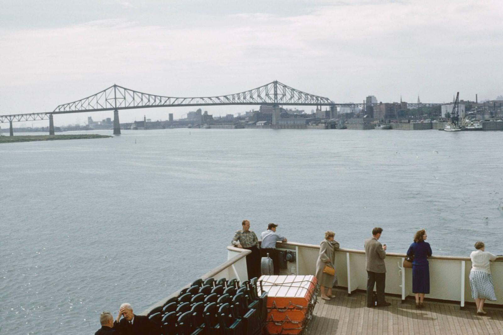 #102 Passengers stand on the deck of a boat on the St Lawrence River beside the port district of the city of Montreal in the province of Quebec in Canada, 1960.