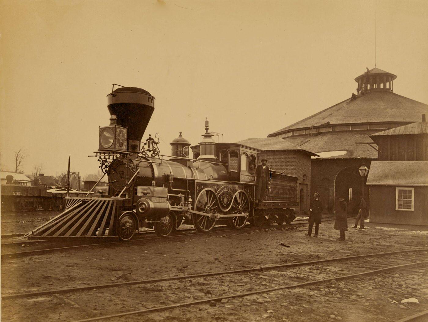 #122 Ornately decorated locomotive J.H. Devereux, of the United States Military Railroad with two crew members on board outside the roundhouse at the Alexandria station, 1863
