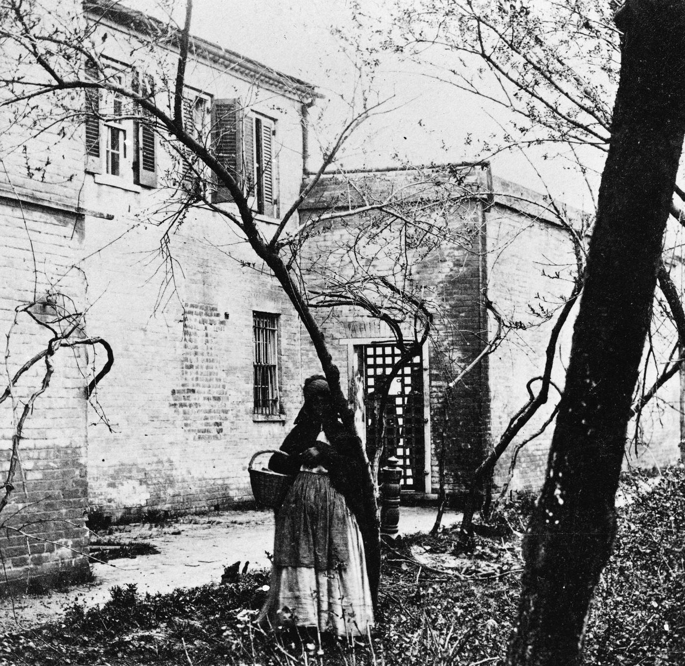 #139 A black woman holds a basket and stands among bare trees in a slave pen, Alexandria, Virginia, 1860s