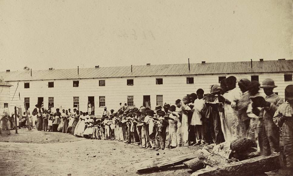 #64 African-American adults and children reading books while lined up in front of a barracks-like building. The same building is shown in “Freedman’s Barracks, Alexandria, 1860s