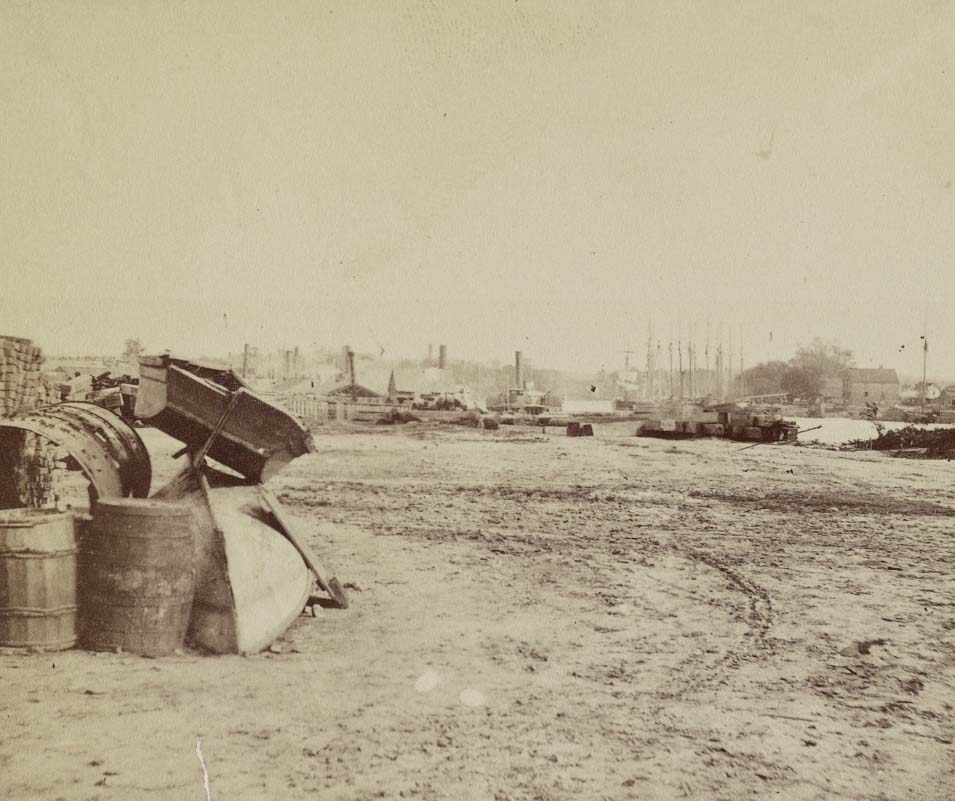 #216 Two boats stacked next to supplies with the wharf in the distance, 1865