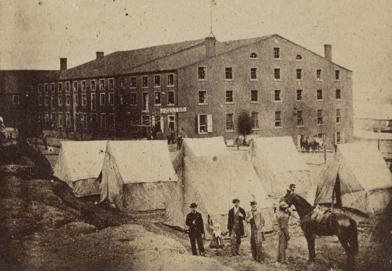 #28 Agroup of people including a child and a man holding on to a horse next to tents in front of Libby Prison, a Confederate military prison established in 1861.