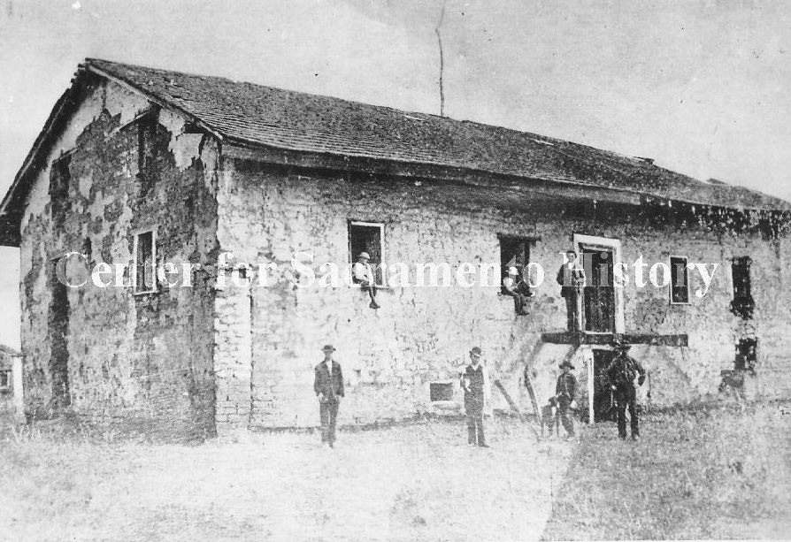 #140 Sutter’s Fort prior to being rebuilt with just the central building left standing. Several men and a dog are sitting in windows and doorways and standing next to the buiding, 1867