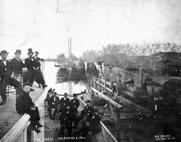#8 The Levee Sacramento 1866. The photo shows boats, railroad tracks with train, and men seated on boat in foreground.