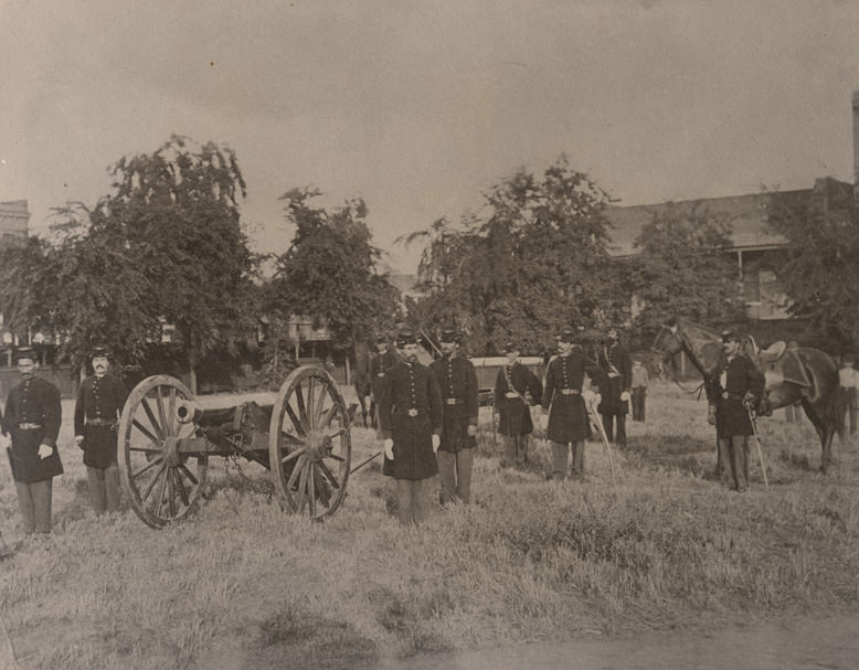 #55 Nine members of the California Milita Batter B. An 1841 Howitzer Cannon at left. The men stand in grassy area, bldgs. at rear, Sacramento City Plaza, 1865