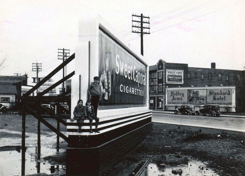 #27 Three young boys are posing on the base of the board. The billboard is opposite the Ridley Arms, a Standard Hotel, at 2301 Danforth Avenue, 1936