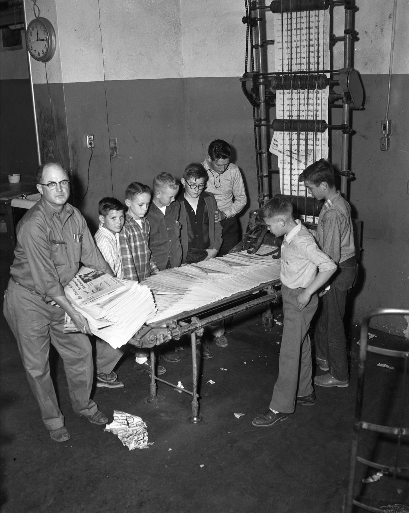 #43 Abilene Reporter News boys and a man standing around a newspaper printer, looking at the stack of newspapers on the machine, 1955