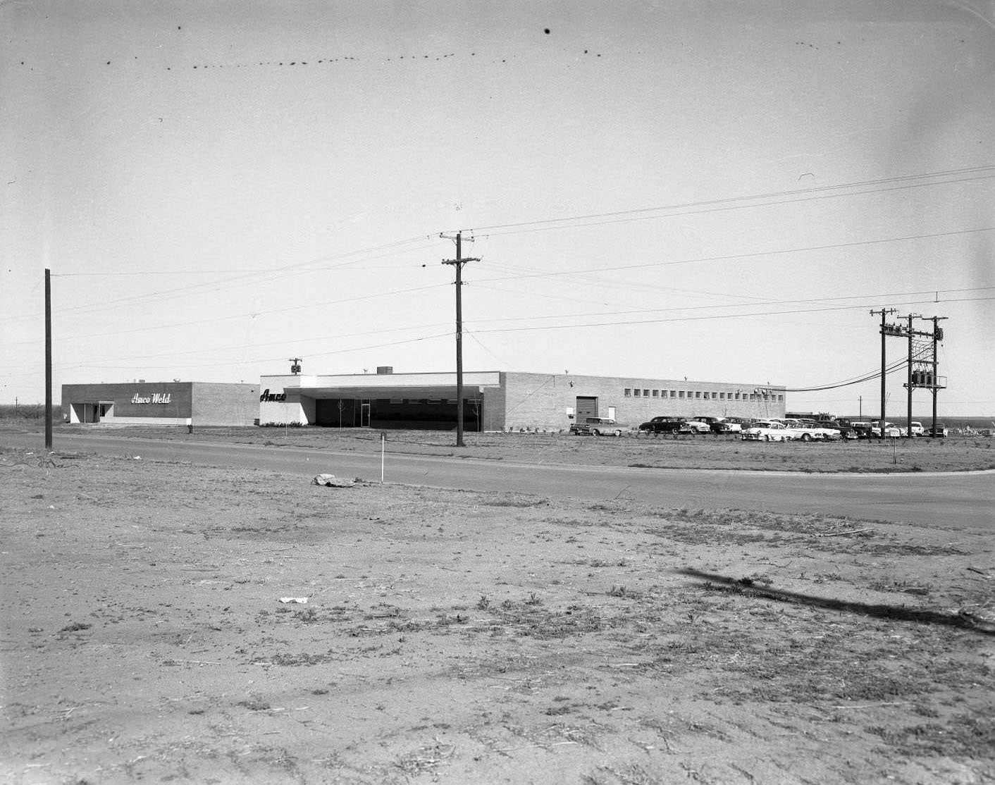 #136 Two Amco buildings on West N. 1st Street in Abilene, 1958. The buildings are viewed from across a street, and there are cars parked beside them and power lines around them.