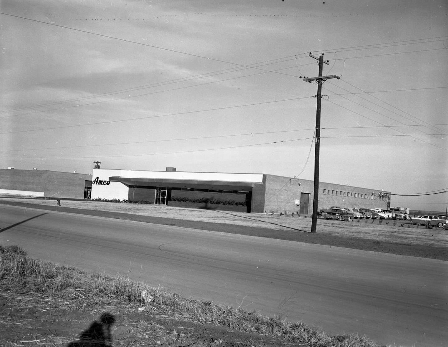 #48 AMCO Manufacturing, 1958. To the side of the building, there are several cars parked in a parking lot. In the foreground, there is a street.