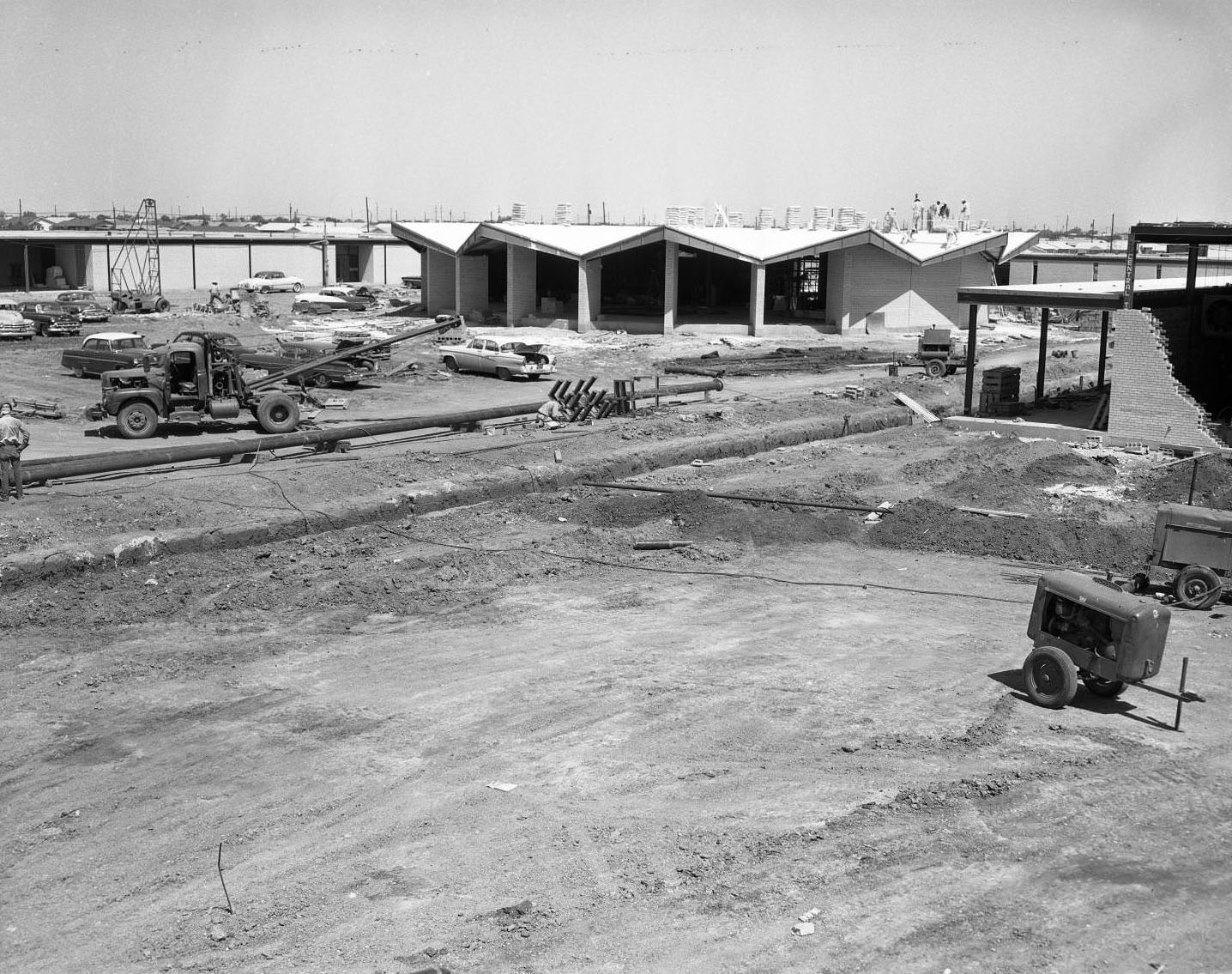 #11 The construction of Cooper High School, 1955. Several buildings are all partially built. There are various pieces of construction machinery around the buildings. Several men stand on top of one of the buildings.