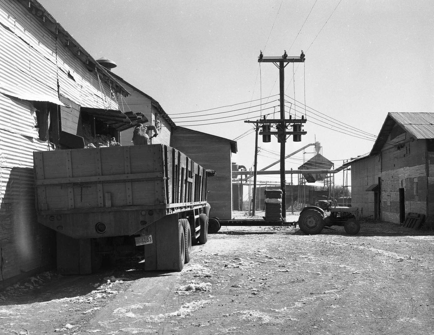 #63 A cotton gin, 1958. There is a truck and trailer being loaded with cotton by a man. There are several buildings and machines with a tractor and power lines between them.