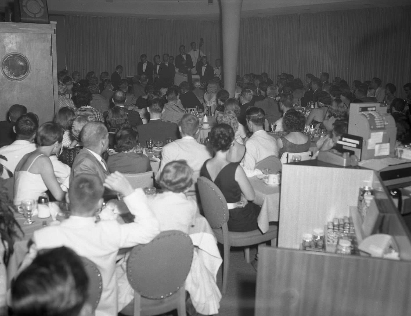 #65 A show at the Sands Hotel in Abilene, 1957. The Crew Cuts quartet is singing on stage at the back of a room with an audience sitting around tables.