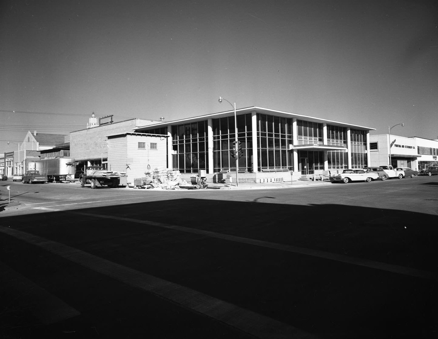 #17 The Abilene Public Library, 1957. The building is on the corner of Cedar and North 2nd Street. There are other buildings on each side of the library, cars parked on the street, and light posts.