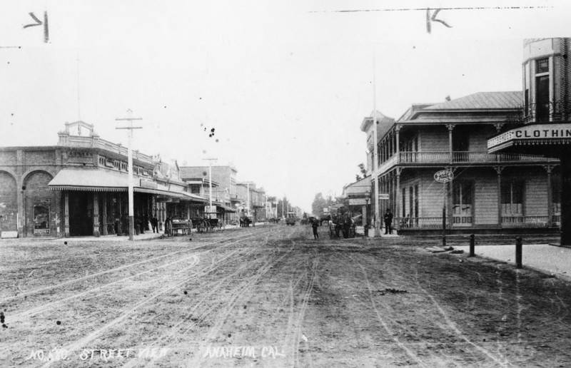 #161 View of an unpaved street in Anaheim in 1890, with shops and stores on either side and carriages parked at curbs. A streetcar is seen on tracks in the middle of the street.