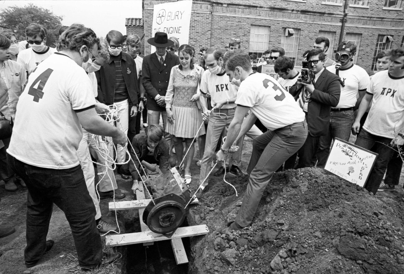 #79 Students at The University of Texas at Arlington gathered around before dropping an internal combustion engine into a shallow grave on campus for Earth Day, 1970