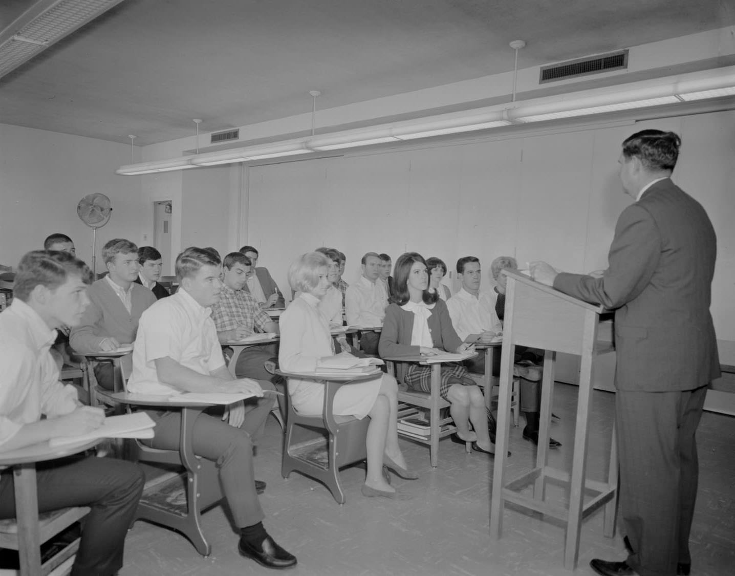 #164 Students sit in desks looking at a man, presumably a lecturer, who is standing behind a wooden podium and speaking in an unknown room at St. Edwards University, 1966