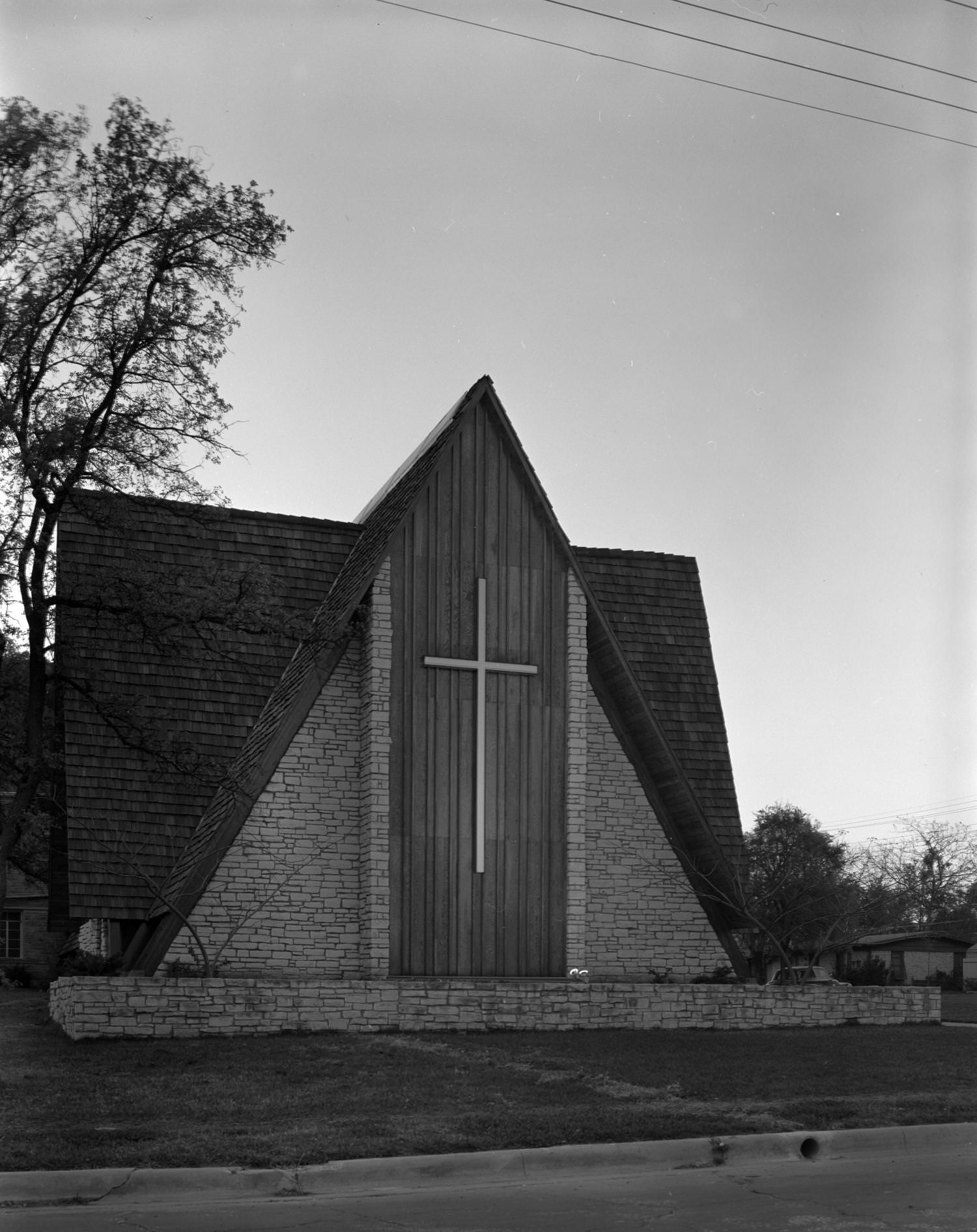#185 A church made from stone, with large, sloping roofs. On the building’s front is a large cross, 1961