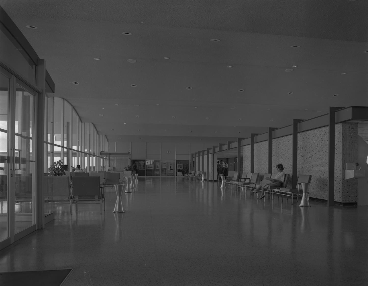 #194 Interior of the Austin Municipal Airport where an open corridor can be seen with seating areas to the left and right, 1961