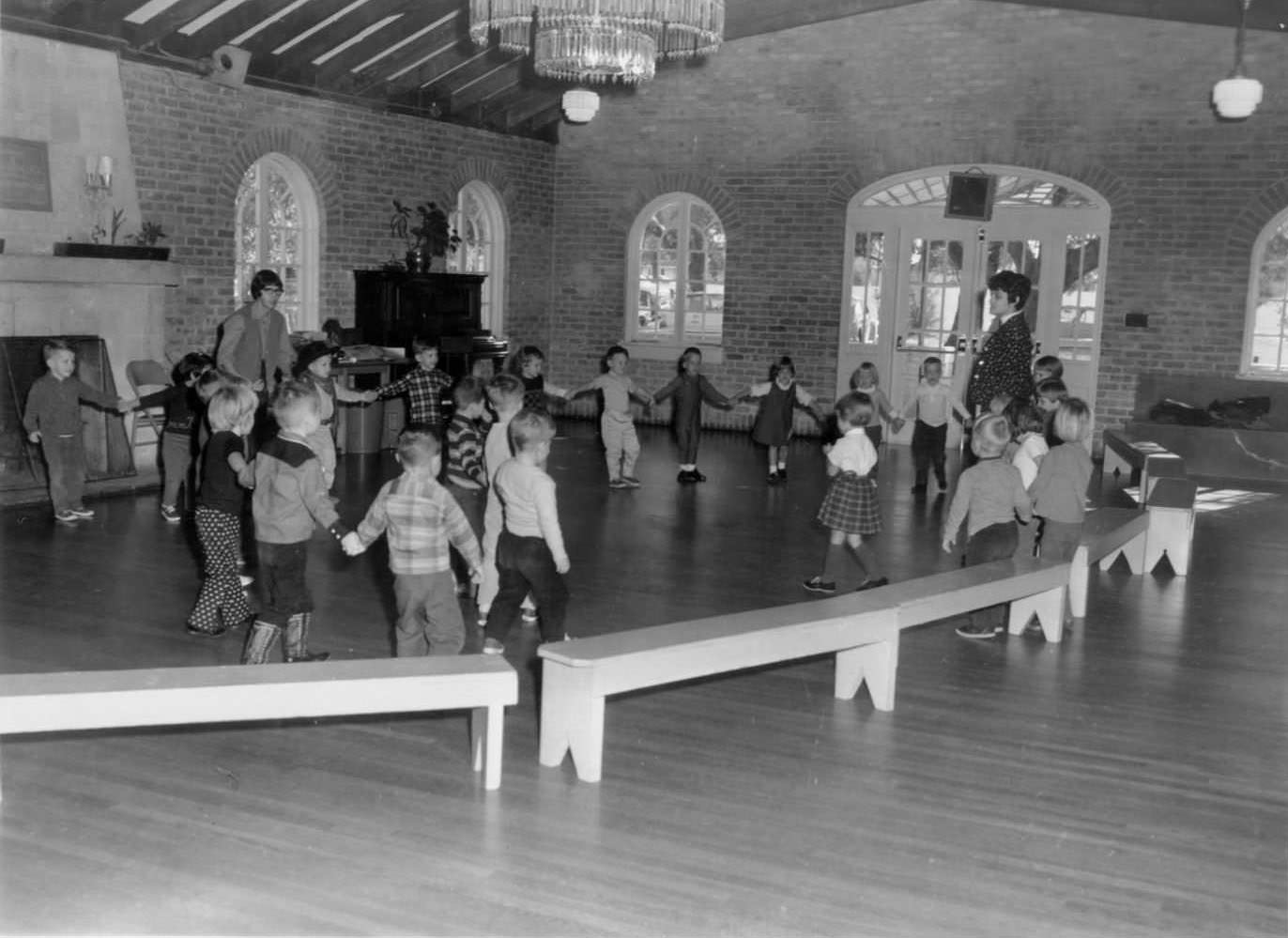 #102 A group of children playing a game. They are in a large circle, holding hands, with two adult instructors, 1966