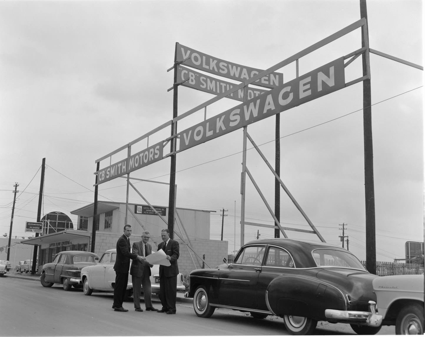 #134 The exterior of CB Smith Volkswagen Motors, Austin, 1960. There are a few cars parked next to the sign advertisement. All three of the men are wearing suits.