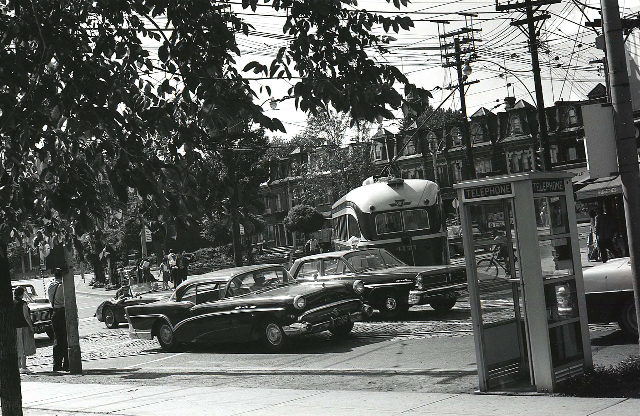 #163 Phone booths, streetcars, and vintage vehicles at Gerrard and Parliament. Taken from the southeast corner, looking northwest across the island parkette in 1963