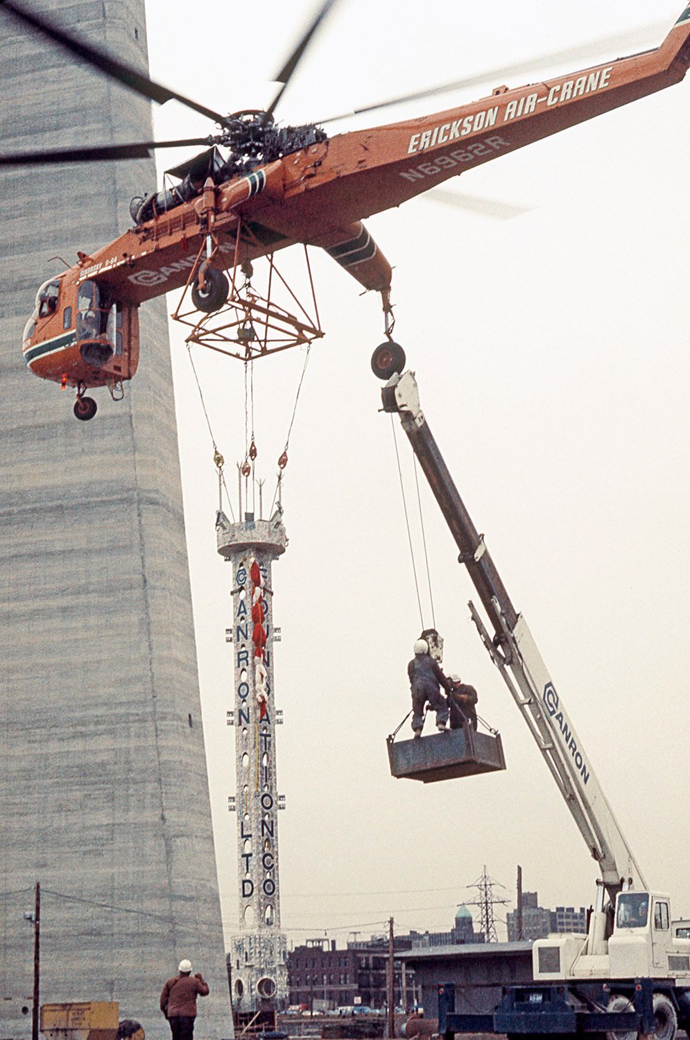 #95 Bob Benns (seen on the ground) managing the last antenna piece lift by Olga the Sikorsky helicopter, 1975
