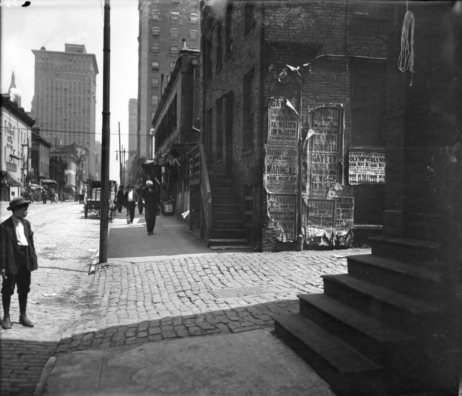 #17 A boy in the foreground is reading an assortment of advertisements plastered to the side of a brick building, 1910