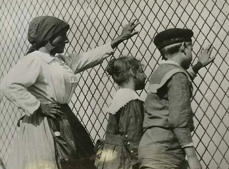 #44 A woman, a boy and a girl at a chain link fence, Ellis Island, New York
