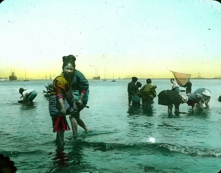 #22 People wading in sea; fisher with hand net; boats in background.