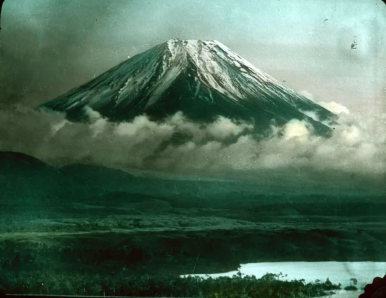 #27 Snow-covered mountain (Mt. Fuji ) wreathed with clouds; low ground leading down to water in foreground.