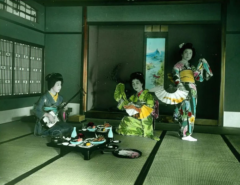 #30 Three women with fans, musical instrument and a table set with food.