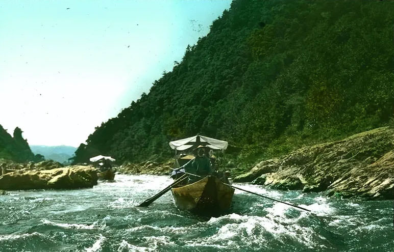 #33 Two boats with cloth canopies being rowed along river; wooded mountain on one side; rushing water in foreground.