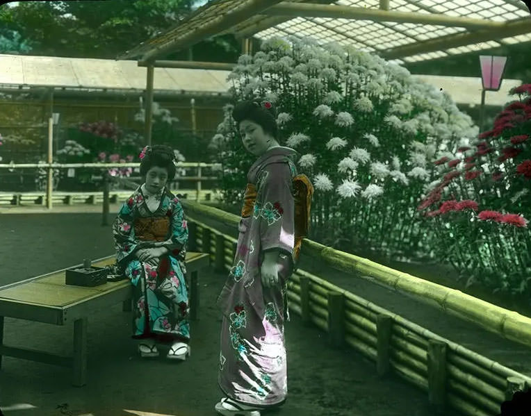 #35 Two young women in Kimonos, one standing, one seated on wooden bench outside open greenhouses filled with blooming flowers.