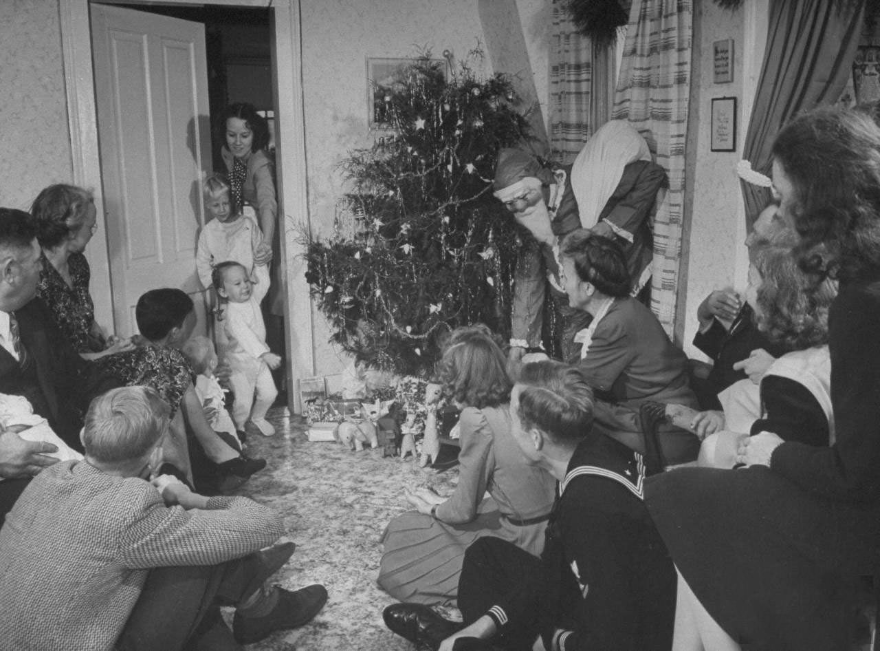 #8 Adult members of farmer James Ferdinand Irwin’s family gathered nr. tree watching his brother-in-law Fred Andrews (in Santa Claus costume) give presents to young family members at early Christmas family reunion marking safe return of sons from service in .WWII