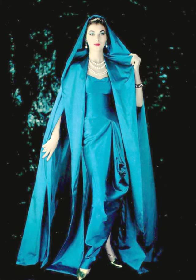 #25 Rose Marie in gorgeous blue draped satin gown gathered at the hip and split at the ankles worn under a hooded cloak of same material by Grès, 1957