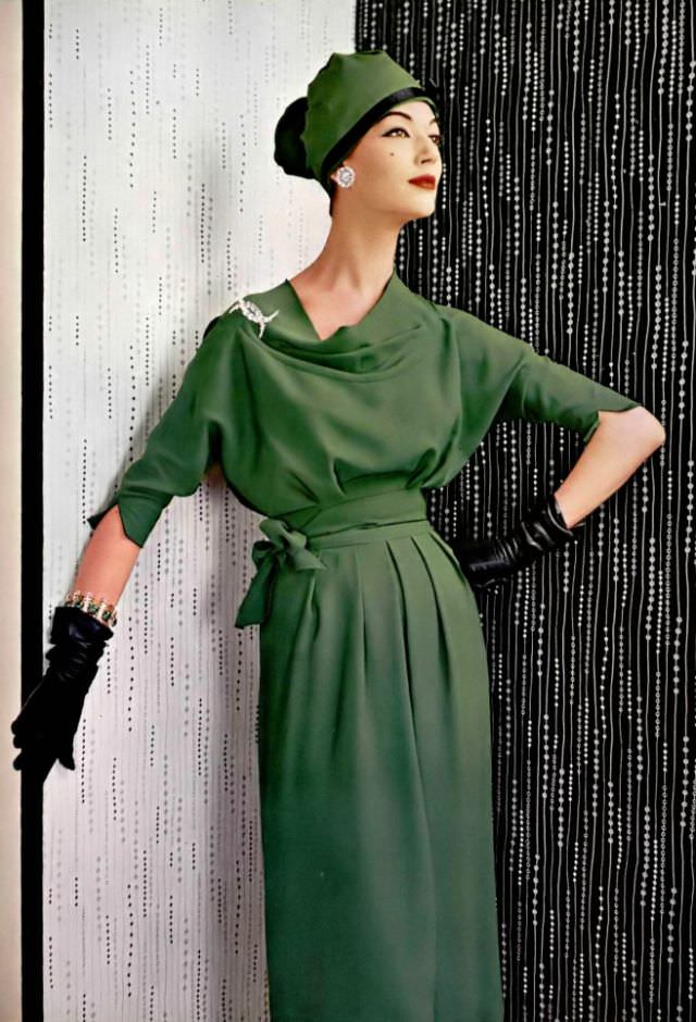 #28 Simone d’Aillencourt in green crêpe dress, the supple bodice with cowl neck ends in scalloped sleeves, belt of the same material is tied in knot at the side, by Grès, jewelry by Roger Scémama, 1957