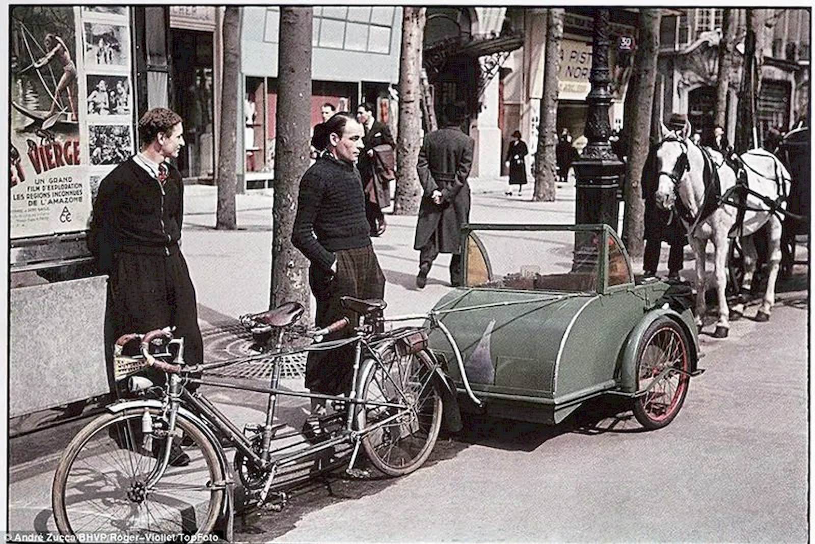 #25 Two fashionably dressed young men stand by a tandem bicycle towing a carriage of sorts.