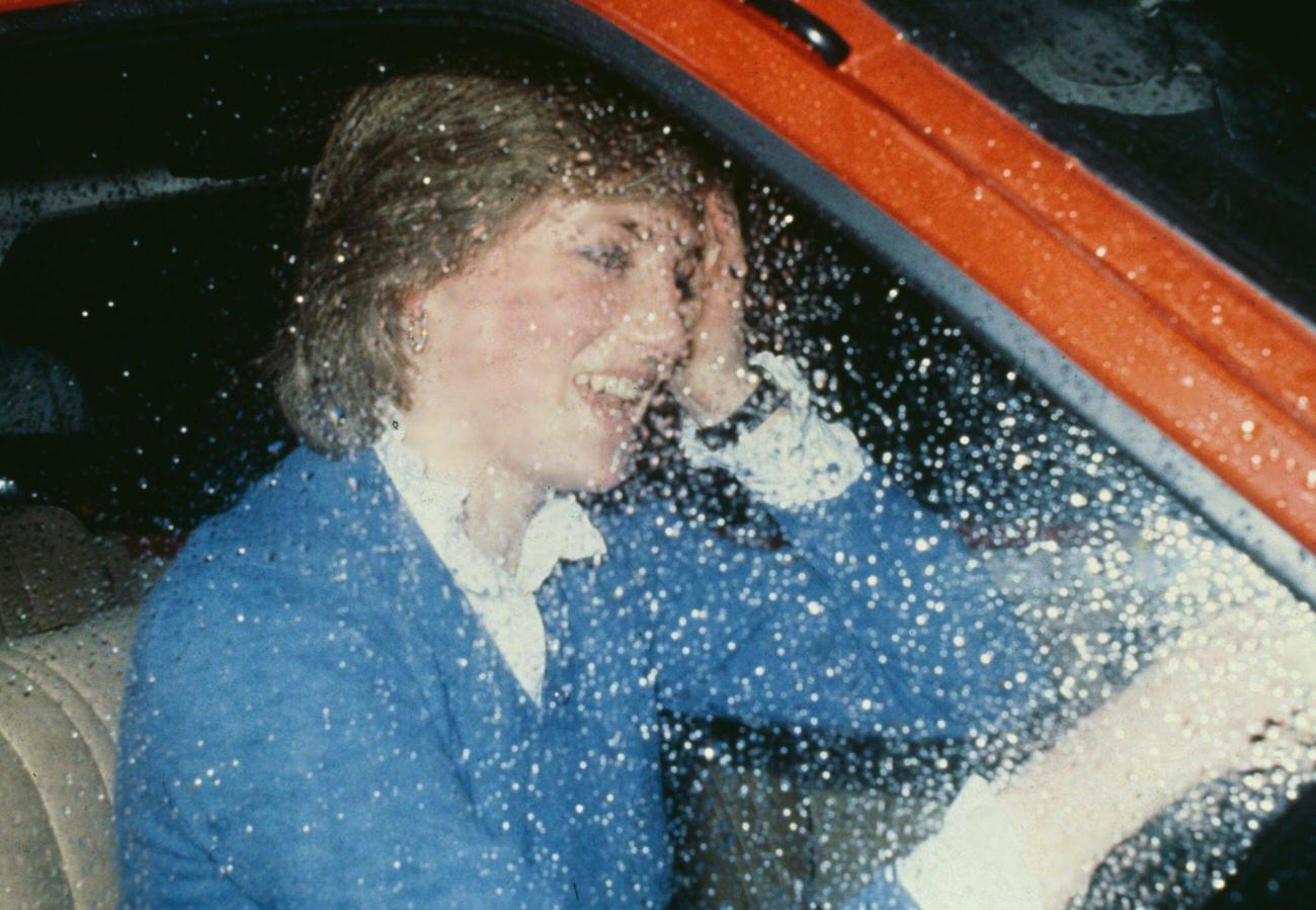 #21 Lady Diana Spencer wearing a blue sweater over a white blouse, rain on the driver’s seat window as she drives her Austin Metro car, 1980