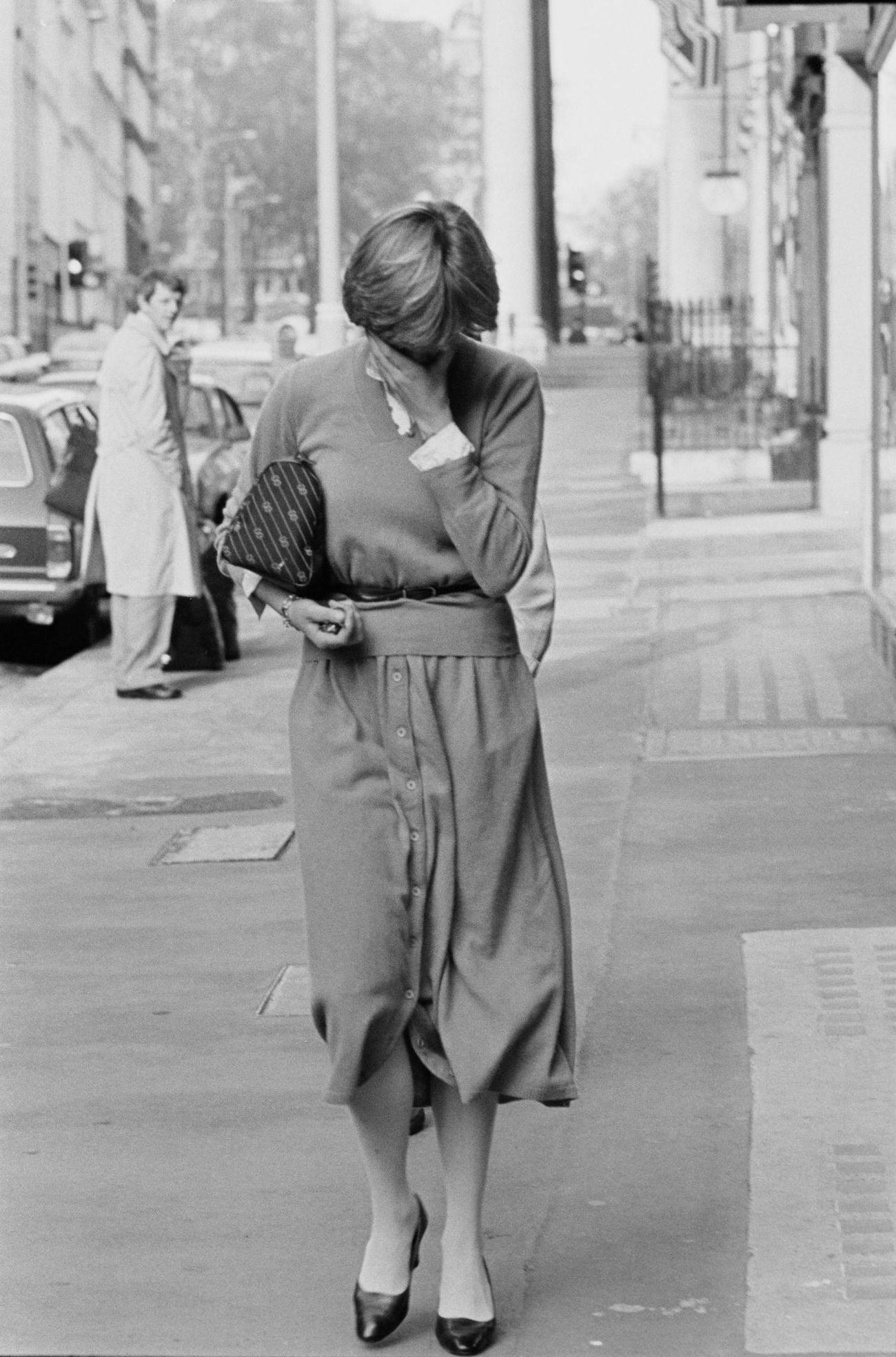 #30 Lady Diana covers her face as she walk on a street in London, UK, 17th November 1980; she is wearing cardigan and midi skirts buttoned on front.