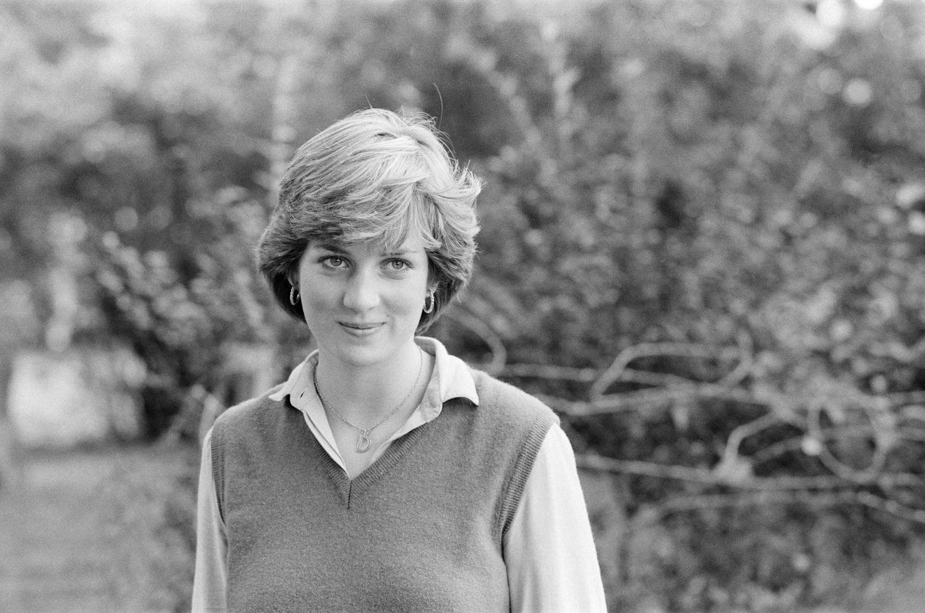 #8 Lady Diana Spencer, wearing a floral pattern dress with a sweater draped over her shoulders, her head bowed with her left hand covering her face to avoid photographers, in St George’s Square.