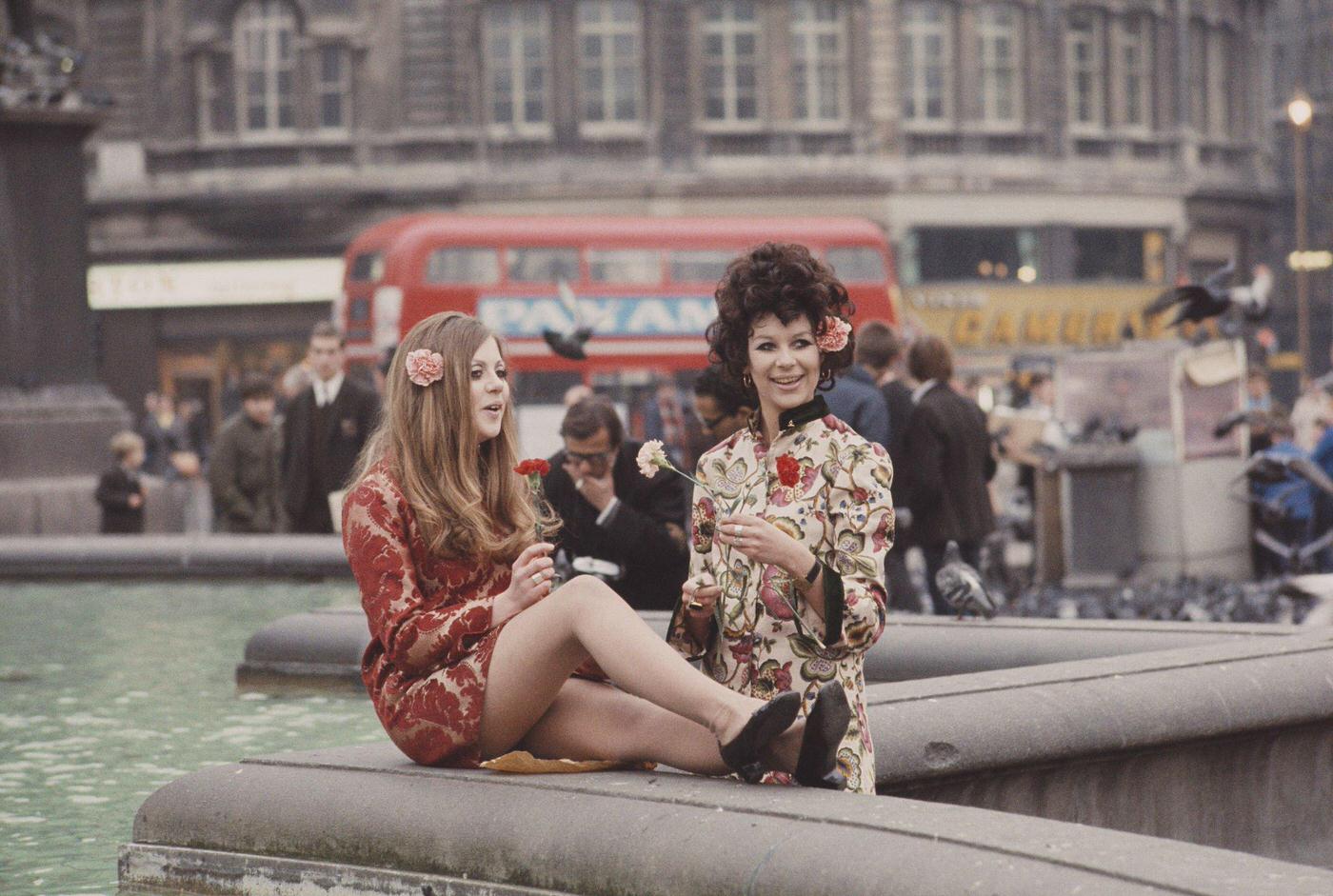 #9 Two young girls dressed in floral print hippie style tunics stand holding flowers beside ornamental fountains in Trafalgar Square, London in November 1967