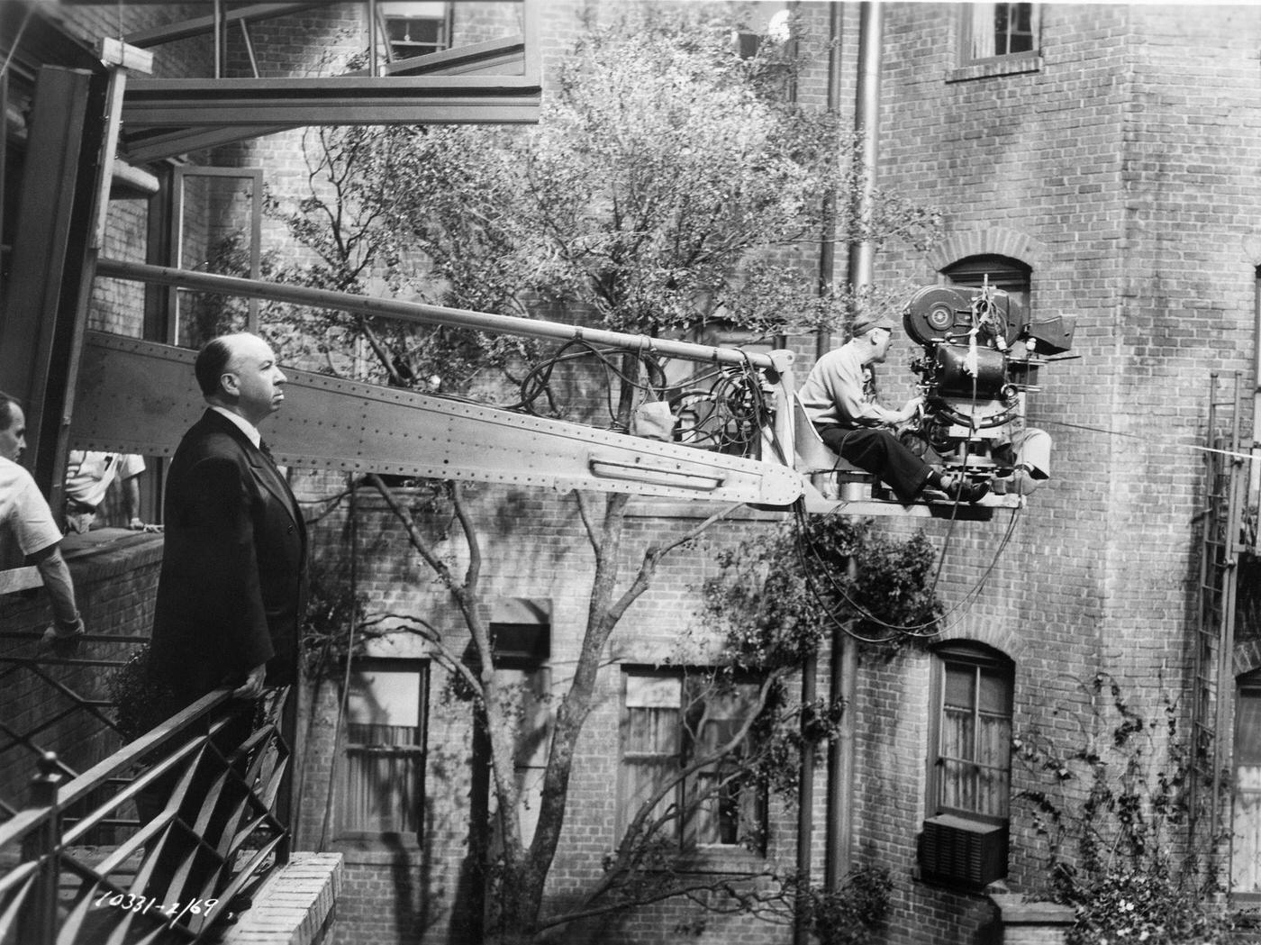 Alfred Hitchcock observes the action from a balcony as cameramen extended out on an “arm” film a scene on the set of “Rear Window” 1954.