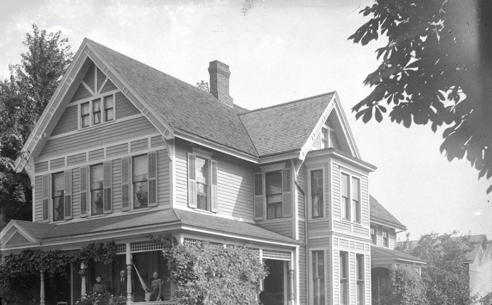 #5 A large house with four people on and around the porch facing the camera, 1900. Going towards the right of the photo, the neighborhood continues with vegetation grown houses.