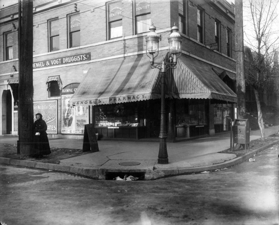 #57 The Mengel & Vogt pharmacy at Taylor and Page Avenues, 1900. A woman is standing next to a pole in front of the building. The building still exists and appears to be vacant.