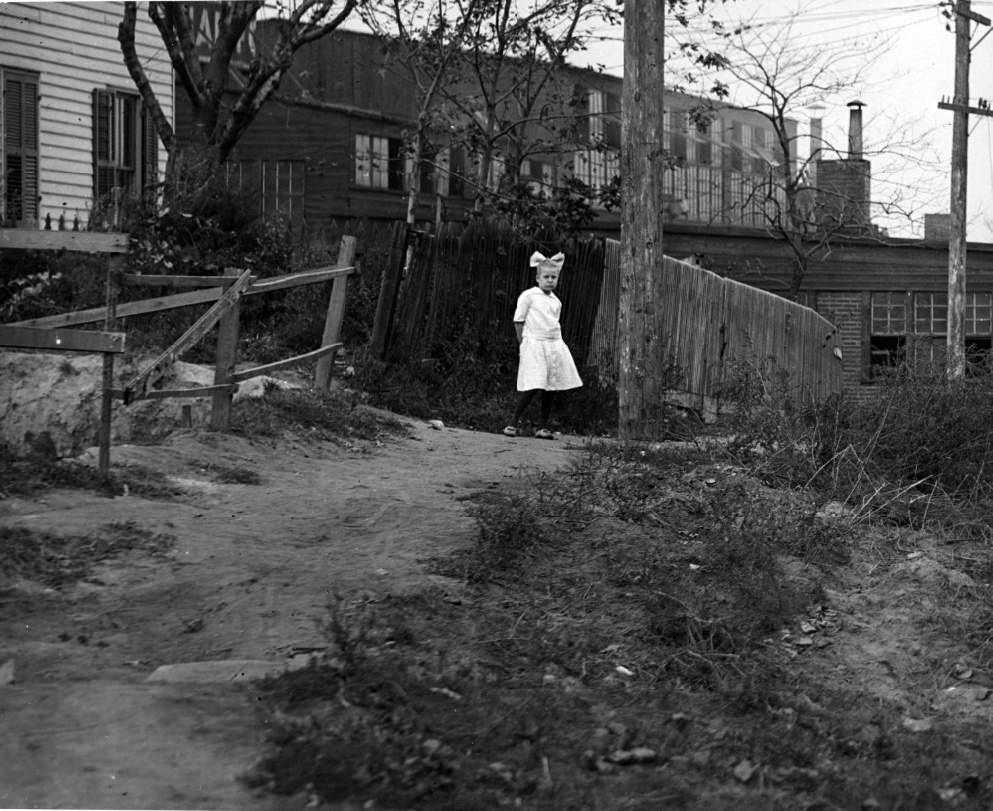 #81 Girl on dirt path, 1900. An industrial warehouse is visible in the background.