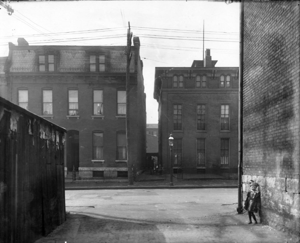 #90 The front of two city houses side by side, 1900. Buildings in foreground partially obstructing the house fronts. Young child in alley in foreground.